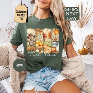 May include: Moss green t-shirt with a fall-themed graphic. The graphic features a scarecrow, pumpkins, and autumn foliage. The words "Fall Vibes" are written in a script font. The shirt is part of the Comfort Colors 1717 collection.
