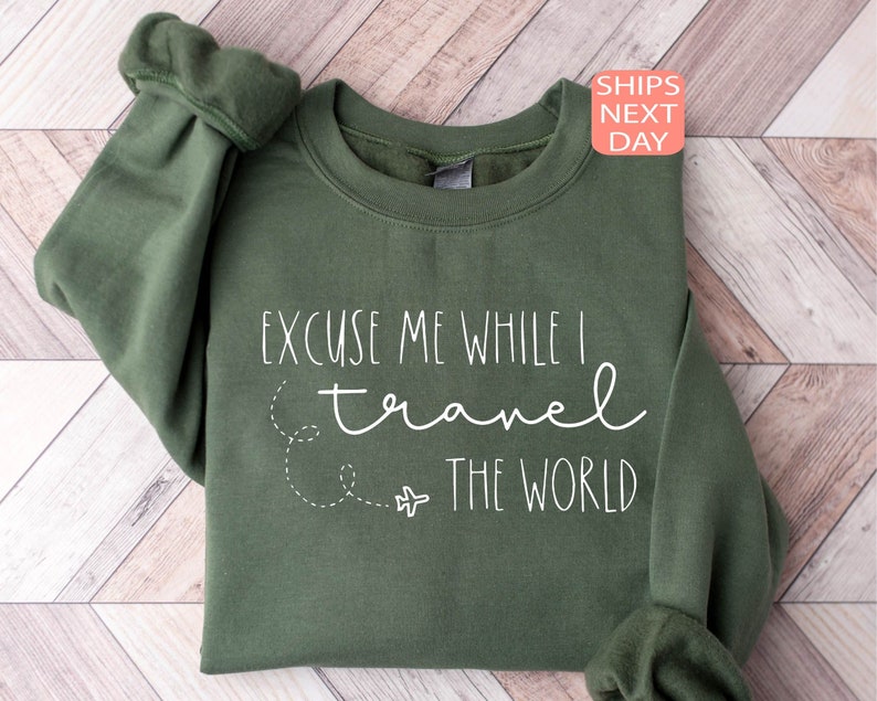 Może przedstawiać: Zielony sweter z napisem "Excuse me while I travel the world" w białym kolorze. Tekst jest uzupełniony małym obrazkiem samolotu i przerywaną linią.