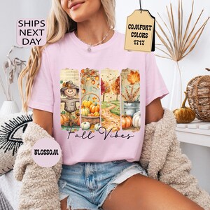 May include: A light pink t-shirt featuring a fall-themed graphic with a scarecrow, pumpkins, and autumn foliage. The words "Fall Vibes" are written in a script font. The shirt has a crew neck and short sleeves.