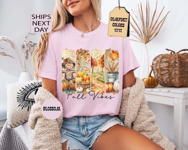 May include: A light pink t-shirt featuring a fall-themed graphic with a scarecrow, pumpkins, and autumn foliage. The words "Fall Vibes" are written in a script font. The shirt has a crew neck and short sleeves.