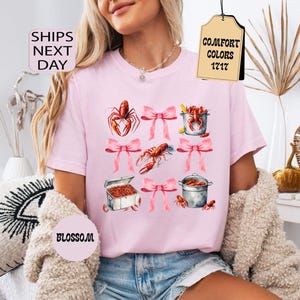 K&ouml;nnte beinhalten: Pinkes T-Shirt mit einem grafischen Design aus Krebsen, T&ouml;pfen und rosafarbenen Schleifen. Auf einem am Shirt befestigten Etikett steht "Comfort Colors 1717".