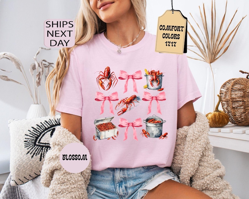 K&ouml;nnte beinhalten: Pinkes T-Shirt mit einem grafischen Design aus Krebsen, T&ouml;pfen und rosafarbenen Schleifen. Auf einem am Shirt befestigten Etikett steht "Comfort Colors 1717".