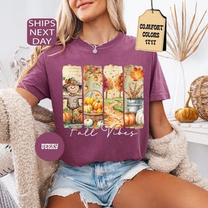 May include: Berry-colored t-shirt with a fall-themed graphic. The graphic features a scarecrow, pumpkins, and autumn foliage. The words "Fall Vibes" are written in a script font. The shirt has short sleeves and a crew neck.