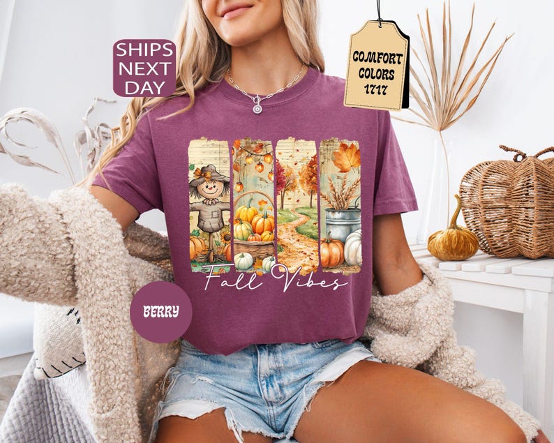 May include: Berry-colored t-shirt with a fall-themed graphic. The graphic features a scarecrow, pumpkins, and autumn foliage. The words "Fall Vibes" are written in a script font. The shirt has short sleeves and a crew neck.