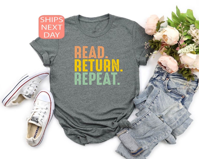 以下が含まれることがあります： オレンジ、黄色、緑で「READ. RETURN. REPEAT.」と書かれた、グレーのTシャツ。白いスニーカーとブルージーンズと組み合わせています。ピンクの花のブーケも含まれています。