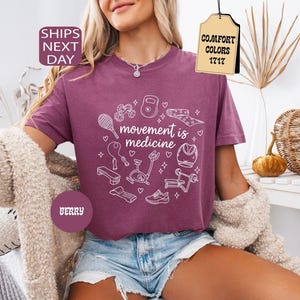 K&ouml;nnte beinhalten: Beerenfarbenes T-Shirt mit dem Schriftzug "movement is medicine" und wei&szlig;en Strichzeichnungen von Trainingsger&auml;ten. Das Shirt hat einen Rundhalsausschnitt und kurze &Auml;rmel. Das Bild enth&auml;lt auch den Text "Comfort Colors 1717".