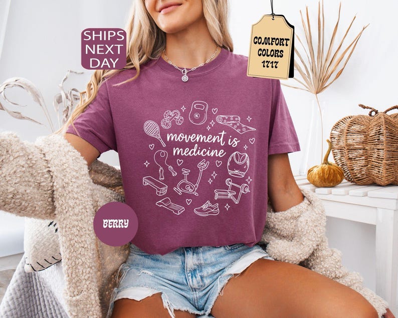 K&ouml;nnte beinhalten: Beerenfarbenes T-Shirt mit dem Schriftzug "movement is medicine" und wei&szlig;en Strichzeichnungen von Trainingsger&auml;ten. Das Shirt hat einen Rundhalsausschnitt und kurze &Auml;rmel. Das Bild enth&auml;lt auch den Text "Comfort Colors 1717".