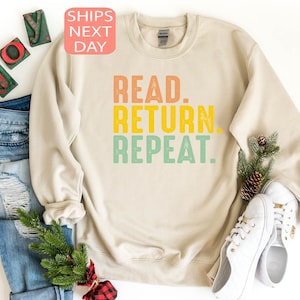 以下が含まれることがあります： オレンジ、黄色、緑で「READ. RETURN. REPEAT.」と書かれたクリーム色のスウェットシャツ。スウェットシャツは、青いジーンズ、白いスニーカー、クリスマスの飾りと一緒に表示されています。