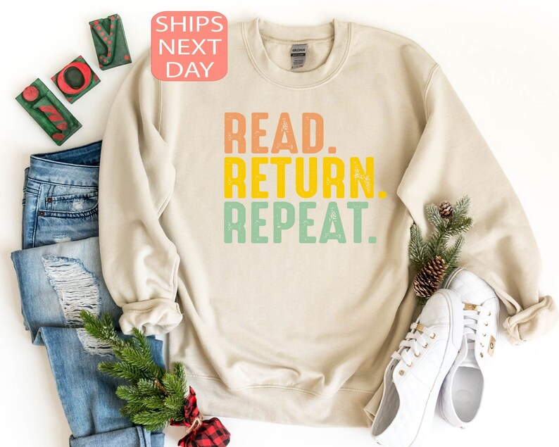 以下が含まれることがあります： オレンジ、黄色、緑で「READ. RETURN. REPEAT.」と書かれたクリーム色のスウェットシャツ。スウェットシャツは、青いジーンズ、白いスニーカー、クリスマスの飾りと一緒に表示されています。