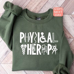 May include: A green sweatshirt with the text "PHYSICAL THERAPY" in white. The text is surrounded by white icons related to physical therapy, such as a dumbbell, a spine, a wheelchair, a cane, a walker, and a person doing a physical therapy exercise.