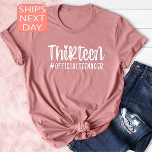 May include: A pink t-shirt with the text "Thirteen #OFFICIALTEENAGER" in white.
