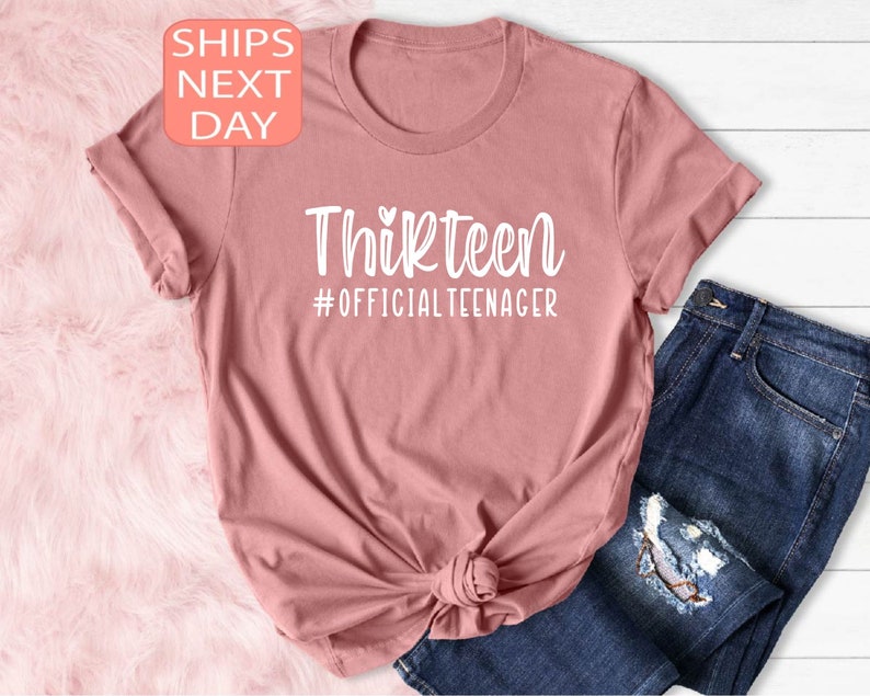 May include: A pink t-shirt with the text "Thirteen #OFFICIALTEENAGER" in white.
