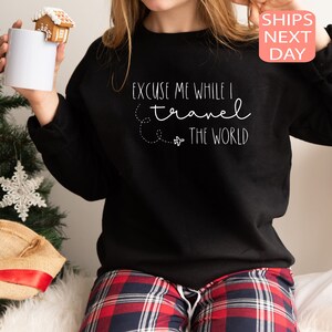 Może przedstawiać: Czarny sweter z białym napisem "Excuse me while I travel the world" i małym nadrukiem samolotu. Model nosi sweter z czerwonymi i czarnymi spodniami w kratę.