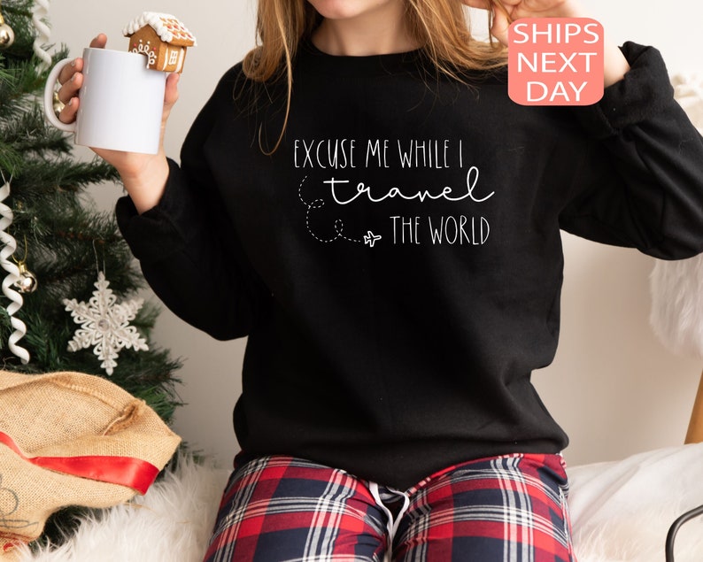 Może przedstawiać: Czarny sweter z białym napisem "Excuse me while I travel the world" i małym nadrukiem samolotu. Model nosi sweter z czerwonymi i czarnymi spodniami w kratę.