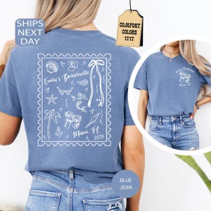 May include: A blue short-sleeved t-shirt featuring a white graphic on the back designed like a postage stamp with illustrations of a mermaid, palm tree, sunglasses, and champagne glasses, with the text Emily's Bachelorette Miami FL 2026.