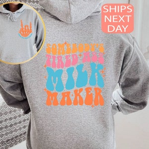 May include: A gray hoodie with the text "SOMEBODY'S TIRED ASS MILK MAKER" in colorful retro font on the back. The front left chest features a skeleton hand graphic with the text "GOOD DAY MILK MAKER".