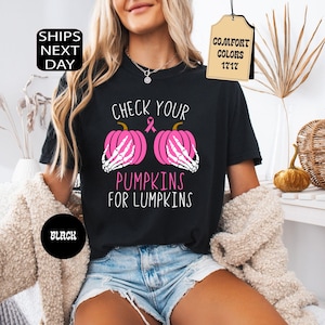 May include: A black t-shirt featuring a graphic of two pink pumpkins held by white skeleton hands, a pink awareness ribbon, and the text "CHECK YOUR PUMPKINS FOR LUMPKINS" in white and pink lettering, with a tag labeled "COMFORT COLORS 1717".