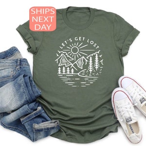 May include: An olive green t-shirt with a white graphic of a mountain scene and the text "LET'S GET LOST". The shirt is paired with blue jeans and white canvas sneakers.
