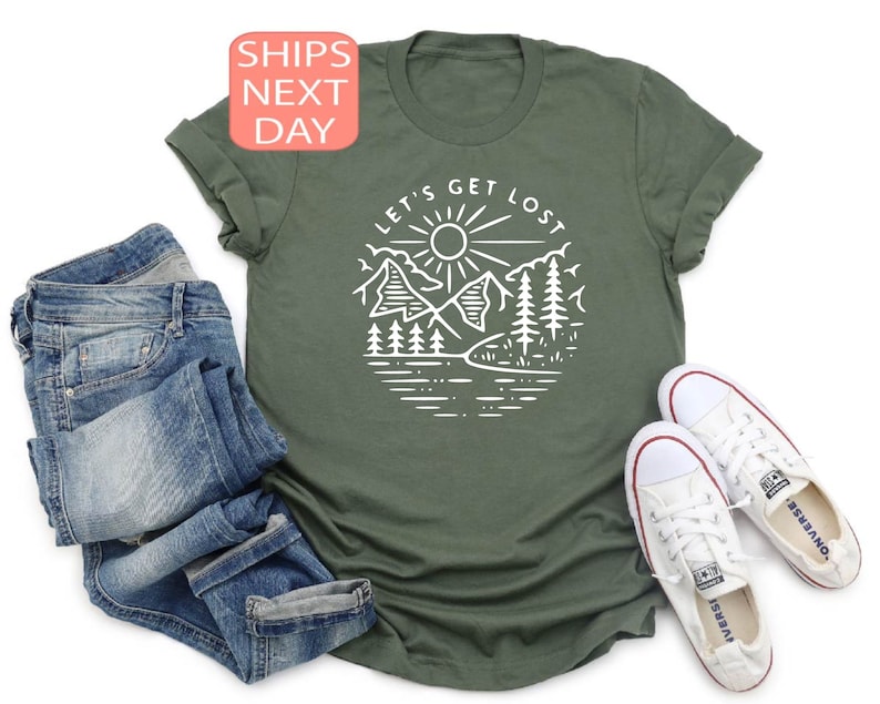 May include: An olive green t-shirt with a white graphic of a mountain scene and the text "LET'S GET LOST". The shirt is paired with blue jeans and white canvas sneakers.