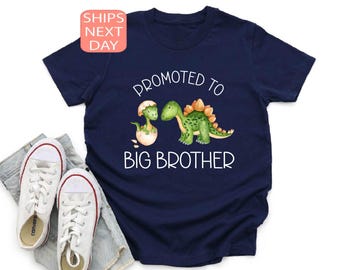 May include: Navy blue t-shirt with the text "Promoted to Big Brother" and a graphic of two green dinosaurs. One dinosaur is hatching from an egg. White sneakers and a pair of jeans are next to the shirt.