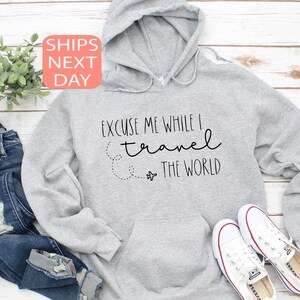 Może przedstawiać: Szara bluza z kapturem z napisem "Przepraszam, podczas gdy podr&oacute;żuję po świecie" i małym rysunkiem samolotu.