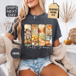 May include: Dark gray t-shirt with a fall-themed graphic. The graphic features a scarecrow, pumpkins, and the text "Fall Vibes." The shirt also has a tag that says "Comfort Colors 1717."