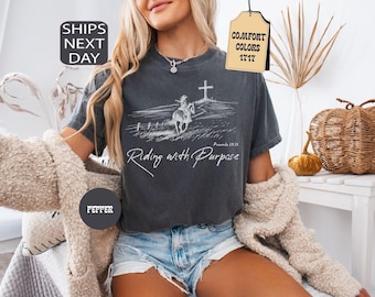 May include: A charcoal gray short-sleeve t-shirt featuring a white line art graphic of a person riding a horse toward a cross on a hill. The text reads Riding with Purpose and Proverbs 19:21. The shirt has a crew neckline and a relaxed fit.