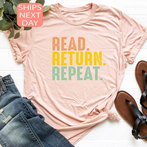 以下が含まれることがあります： オレンジ、黄色、緑色の「READ. RETURN. REPEAT.」という文字がプリントされた、薄いピンク色のTシャツ。青いジーンズと茶色のサンダルも写っています。テキストは太字のレトロなフォントです。