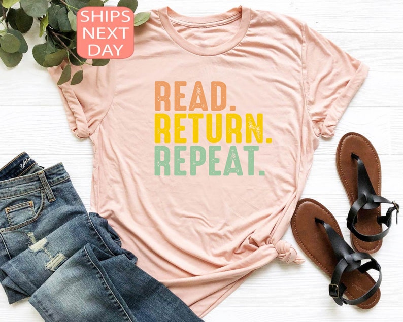 以下が含まれることがあります： オレンジ、黄色、緑色の「READ. RETURN. REPEAT.」という文字がプリントされた、薄いピンク色のTシャツ。青いジーンズと茶色のサンダルも写っています。テキストは太字のレトロなフォントです。