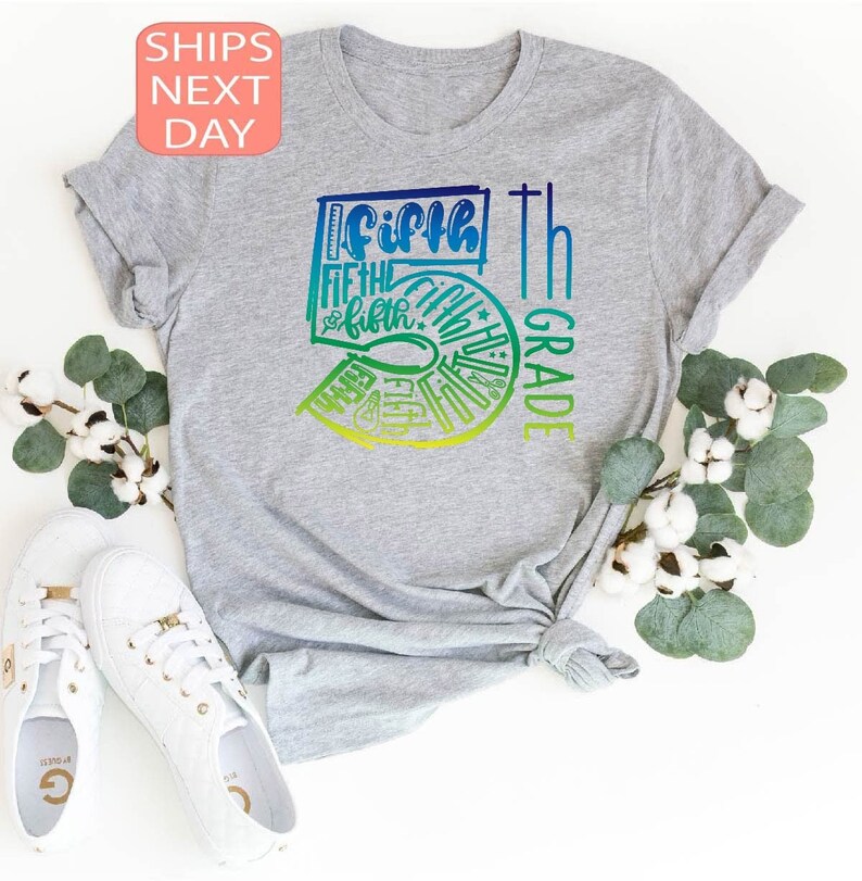 Pu&ograve; includere: Una maglietta grigia con una grafica colorata che dice "Fifth Grade". Il design presenta la parola "Fifth" in un gradiente blu e verde, con il numero "5" in un gradiente simile. La maglietta &egrave; abbinata a scarpe da ginnastica bianche e rametti di cotone.