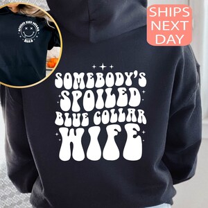 以下が含まれることがあります： 白い文字で「Somebody's Spoiled Blue Collar Wife」と書いてある黒いフード付きスウェットシャツ。レトロなフォントで書かれており、星で囲まれています。
