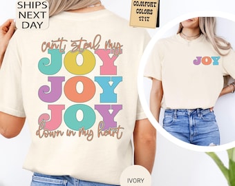 May include: An ivory short-sleeve t-shirt featuring a multi-colored graphic on the back with the text "can't steal my JOY JOY JOY down in my heart" in retro-style lettering. A smaller "JOY" graphic is printed on the front left chest area.