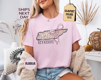 Könnte beinhalten: Ein hellrosa T-Shirt mit einer Grafik des Bundesstaates Tennessee, die mit einem bunten Blumenmuster gefüllt und mit einer rosa Schleife verziert ist. Das Wort Tennessee ist in schwarzer Schreibschrift unter dem Umriss des Staates aufgedruckt.