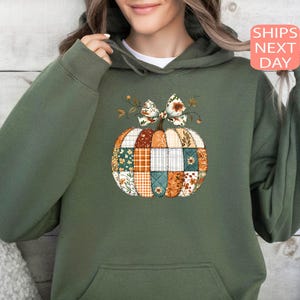 May include: Green hoodie featuring a patchwork pumpkin design with a floral bow. The pumpkin is composed of various fabric patterns in orange, white, and teal. The hoodie has a front pocket and a drawstring hood.