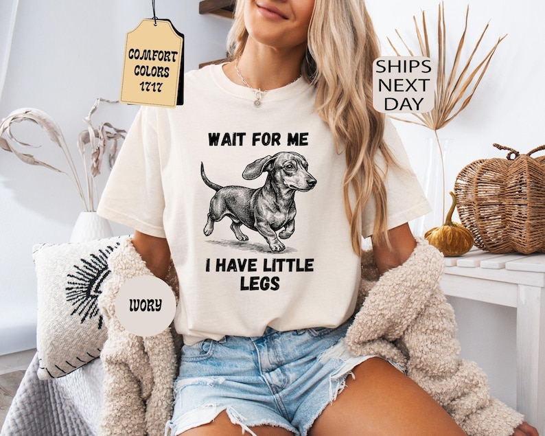 May include: Ivory-colored t-shirt featuring a dachshund illustration with the text "WAIT FOR ME I HAVE LITTLE LEGS." The shirt has a classic crew neck and short sleeves. The image also includes tags that say "Comfort Colors 1717" and "Ivory."