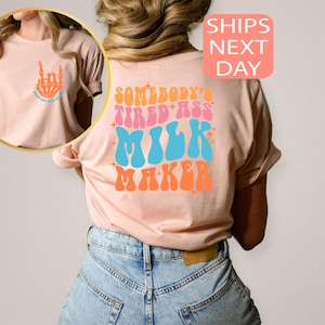 May include: A light peach-colored t-shirt with a retro design. The front features a skeleton hand with the text "Feed All The Babies". The back of the shirt displays the words "Somebody's Tired Ass Milk Maker" in a colorful, groovy font.