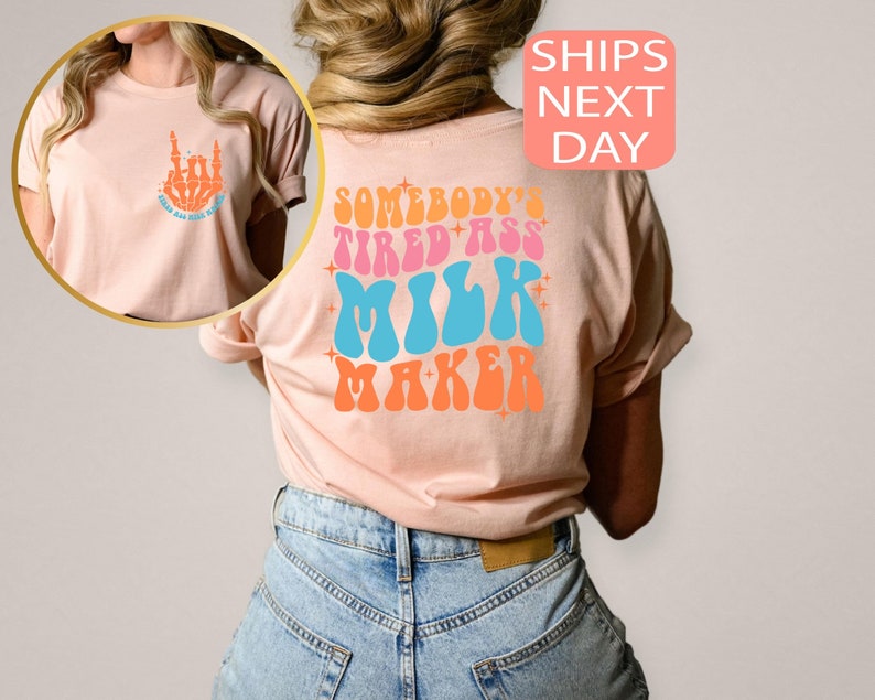 May include: A light peach-colored t-shirt with a retro design. The front features a skeleton hand with the text "Feed All The Babies". The back of the shirt displays the words "Somebody's Tired Ass Milk Maker" in a colorful, groovy font.