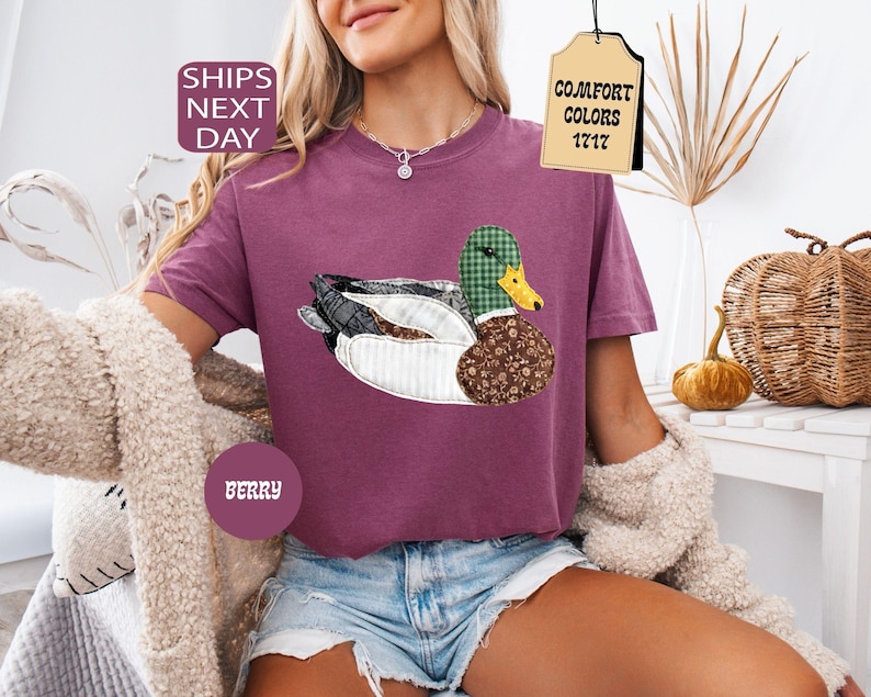 May include: Berry-colored t-shirt featuring a duck applique with plaid, floral, and white fabric details. The duck has a green plaid head and a yellow beak. The shirt has a relaxed fit and is paired with denim shorts.