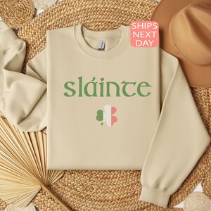 May include: A beige sweatshirt with the Irish word "sl&aacute;inte" printed in green on the front. The word is surrounded by a green shamrock with a pink and white center.