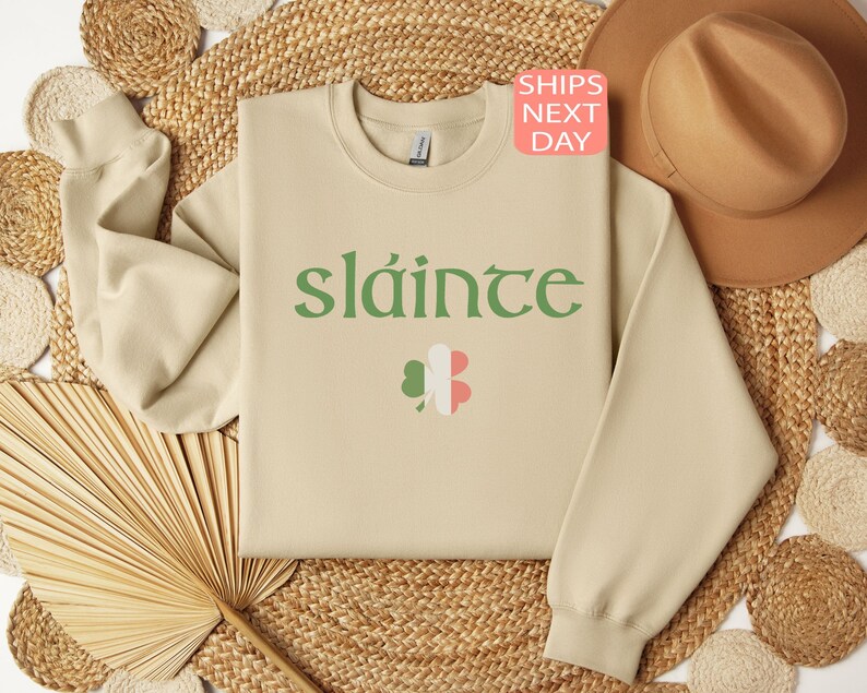 May include: A beige sweatshirt with the Irish word "sl&aacute;inte" printed in green on the front. The word is surrounded by a green shamrock with a pink and white center.
