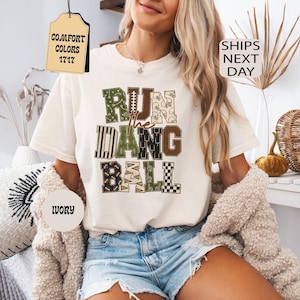 以下が含まれることがあります： アイボリー色の半袖Tシャツで、RUN THE DANG BALLという文字が大きくブロック体でプリントされています。各文字は、フットボールやヘルメットなど、緑、茶色、クリーム色のフットボールをテーマにした様々なパターンで埋め尽くされています。