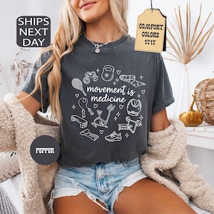 K&ouml;nnte beinhalten: Dunkelgraues T-Shirt mit dem Text "movement is medicine", umgeben von wei&szlig;en Strichzeichnungen von Fitnessger&auml;ten. Das Shirt hat einen Rundhalsausschnitt und kurze &Auml;rmel. Das Wort "Pepper" ist auf einem dunkelgrauen Kreis aufgedruckt.