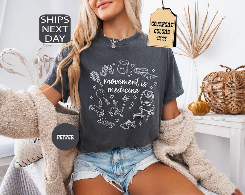 K&ouml;nnte beinhalten: Dunkelgraues T-Shirt mit dem Text "movement is medicine", umgeben von wei&szlig;en Strichzeichnungen von Fitnessger&auml;ten. Das Shirt hat einen Rundhalsausschnitt und kurze &Auml;rmel. Das Wort "Pepper" ist auf einem dunkelgrauen Kreis aufgedruckt.