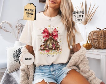 以下が含まれることがあります： アイボリーの半袖Tシャツ。金色の星が散りばめられた大きな赤いリボンの下にキリスト降誕のシーンが描かれています。テキストには「the greatest gift of all」と「Matthew 1:21」と記されています。シャツには「COMFORT COLORS 1717」と「IVORY」のタグが付いています。