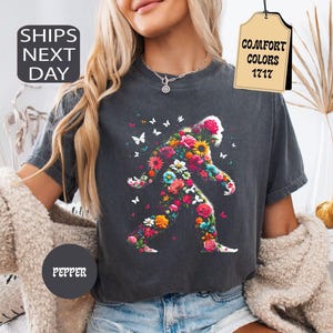 May include: A charcoal grey short-sleeve t-shirt featuring a graphic of a Bigfoot silhouette filled with colourful flowers and butterflies. The shirt has a crew neckline and a relaxed fit. The text COMFORT COLORS 1717 is visible on a tag.