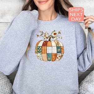 May include: A light gray sweatshirt featuring a patchwork pumpkin design. The pumpkin is composed of various fabric patterns in orange, green, and brown, with a decorative bow and the text "SHIPS NEXT DAY".