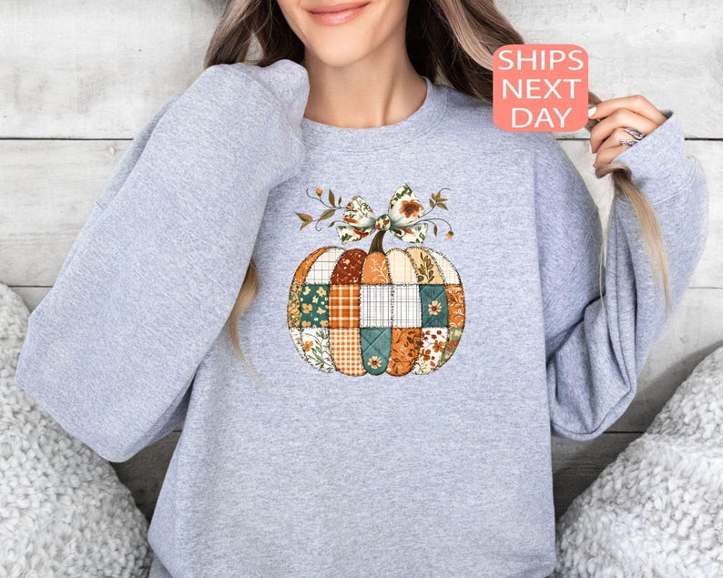 May include: A light gray sweatshirt featuring a patchwork pumpkin design. The pumpkin is composed of various fabric patterns in orange, green, and brown, with a decorative bow and the text "SHIPS NEXT DAY".