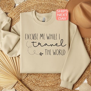Może przedstawiać: Beżowa bluza z napisem "Excuse me while I travel the world" czarnymi literami. Obok tekstu narysowano mały obrazek samolotu.