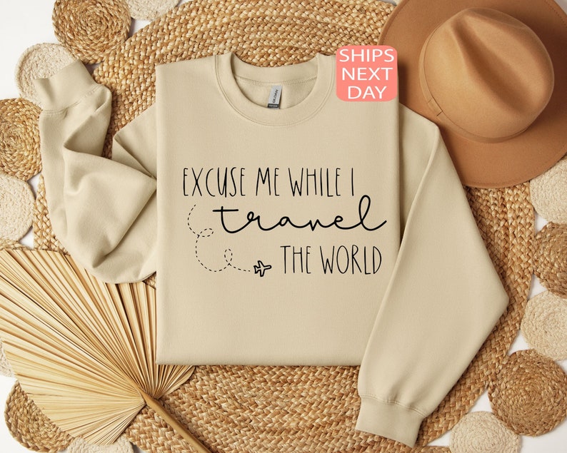 Może przedstawiać: Beżowa bluza z napisem "Excuse me while I travel the world" czarnymi literami. Obok tekstu narysowano mały obrazek samolotu.