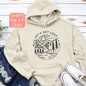 May include: A beige hoodie with a black graphic design featuring mountains, trees, a sun, and the text "LET'S GET LOST". The hoodie is paired with blue jeans and white sneakers. The image is set against a light wood background.